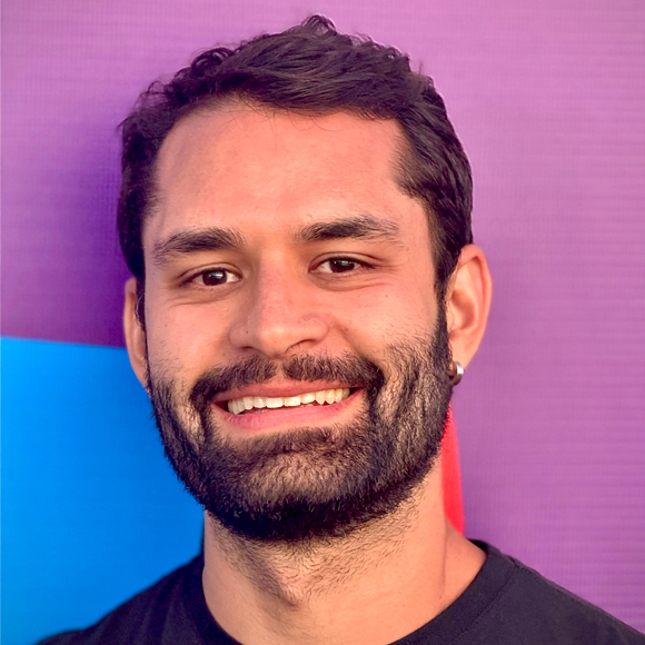 Homem de barba cheia e cabelo curto castanho, sorrindo, usando camiseta preta, em frente a fundo roxo e azul