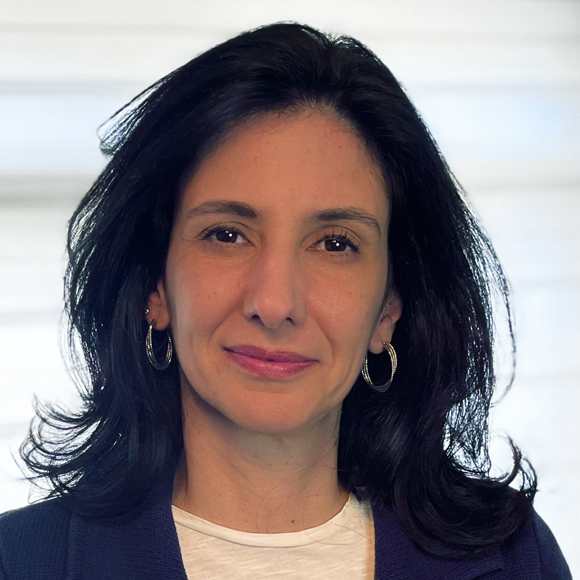 Mulher de cabelos castanhos longos, usando brincos de argola, blusa branca e casaco azul escuro, em frente a fundo claro