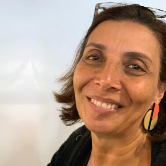 Mulher de cabelos castanhos médios, usando brinco grande colorido e óculos apoiados na cabeça, sorrindo em frente a fundo branco