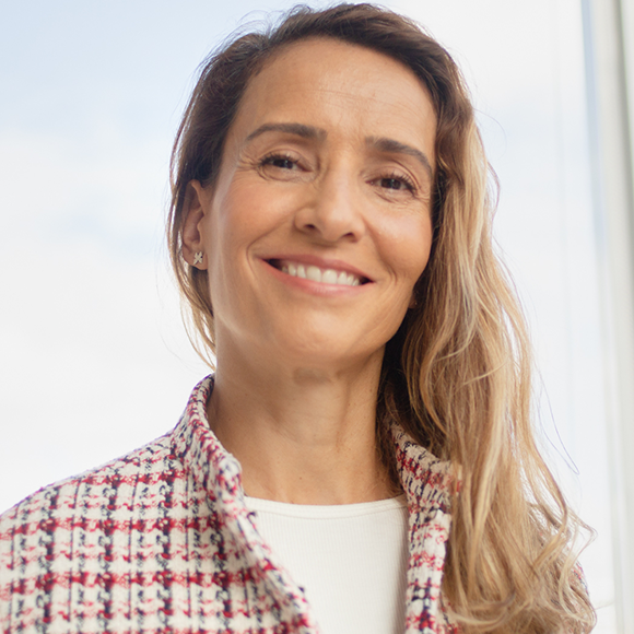 Mulher sorridente de cabelos loiros, usando casaco estampado em rosa e branco, em frente a uma janela iluminada