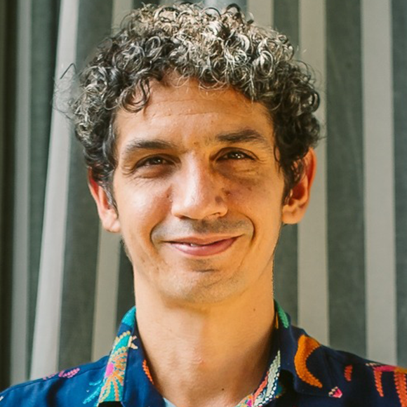 Homem de cabelos cacheados curtos e grisalhos, sorrindo, usando camisa estampada colorida, em frente a cortina listrada