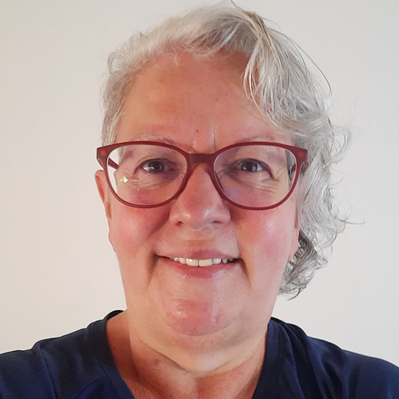 Mulher de cabelos grisalhos curtos, usando óculos vermelhos e camiseta azul escura, sorrindo