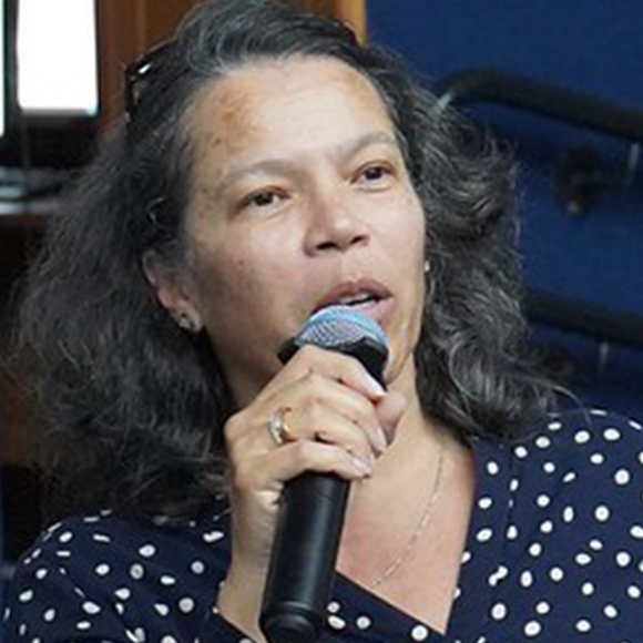 Mulher de cabelos médios e ondulados, usando blusa azul marinho de bolinhas brancas, falando ao microfone em ambiente interno.