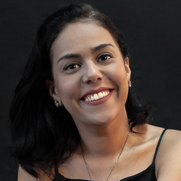 Mulher de cabelos pretos, médios e lisos, vestindo blusa preta de alças finas, sorrindo em frente a um fundo preto.