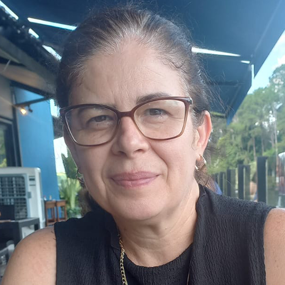 Mulher de óculos, cabelo preso, usando blusa preta, sorrindo levemente em ambiente externo com toldo e árvores ao fundo