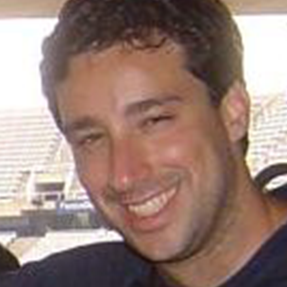 Homem de cabelos curtos e cacheados, sorrindo, em ambiente iluminado com arquibancadas desfocadas ao fundo.