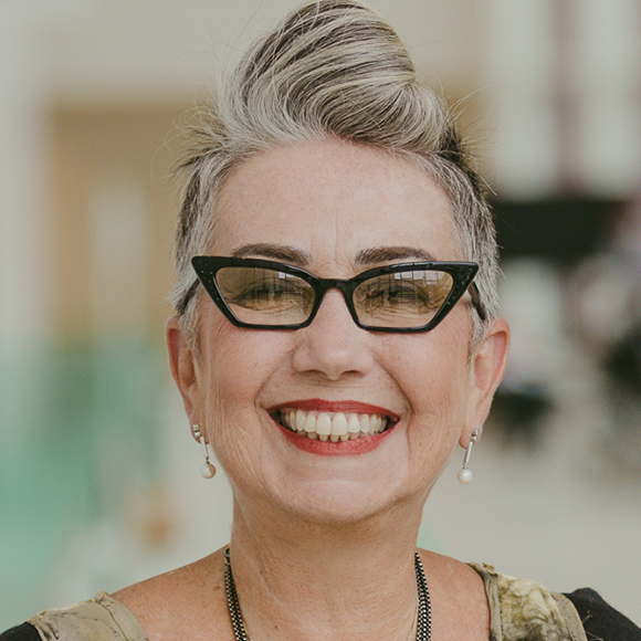 Mulher de cabelos curtos grisalhos, usando óculos de armação preta, brincos de pérola e blusa estampada, sorrindo em ambiente interno iluminado.