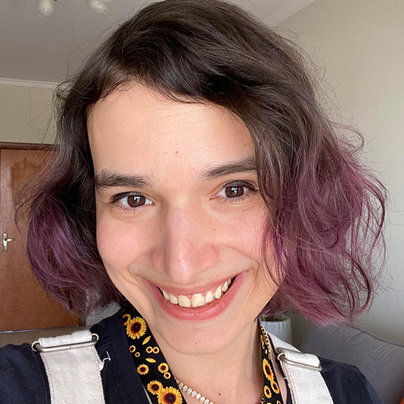 Pessoa sorridente de cabelos curtos castanhos com pontas roxas, usando camiseta preta e jardineira clara, em ambiente interno iluminado