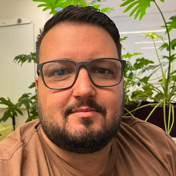 Homem de óculos, barba curta e camiseta marrom, olhando para a câmera em um ambiente interno com plantas verdes ao fundo.