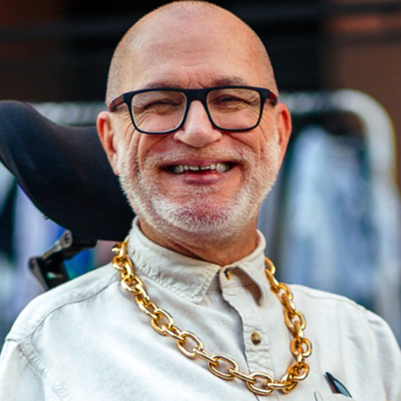 omem sorridente, calvo e de barba grisalha curta, usando óculos escuros de grau, camisa clara e corrente dourada grossa, sentado em cadeira de rodas